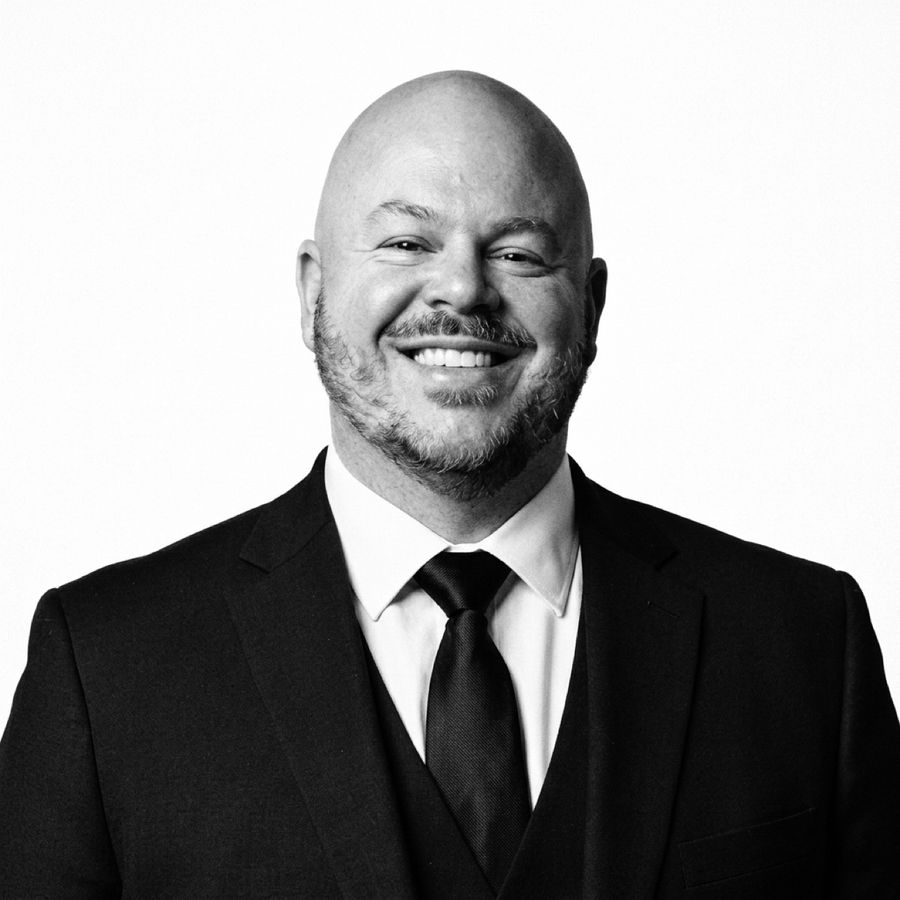 Black-and-white portrait of Sean Anger smiling, wearing a dark suit, vest and tie.